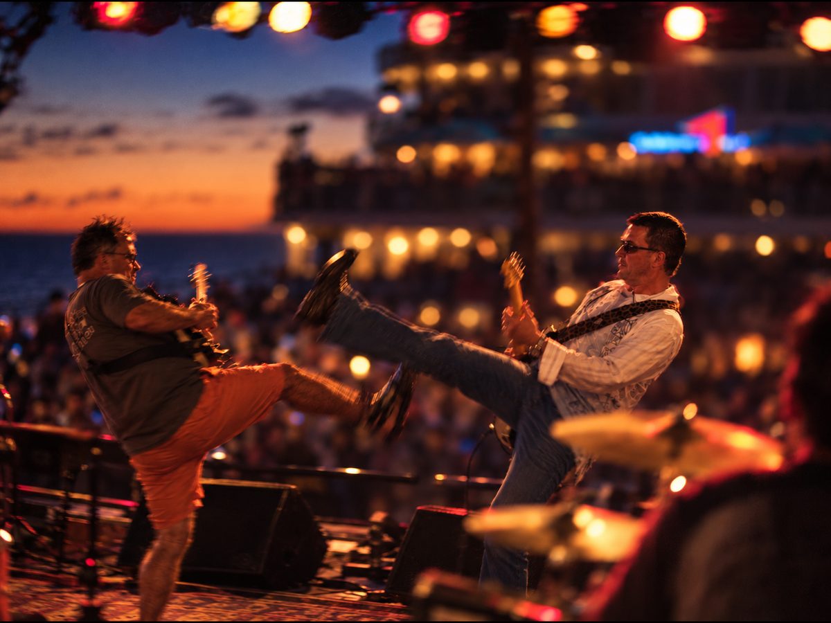 Two guitarists from Men Without Youth trading high kicks on an outdoor stage at sunset